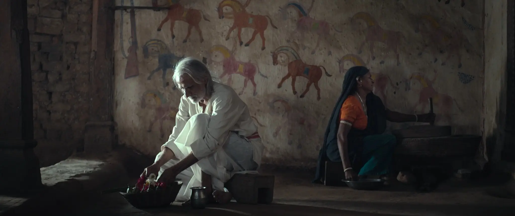 An elderly man and a woman seated apart in a room painted with folk horses